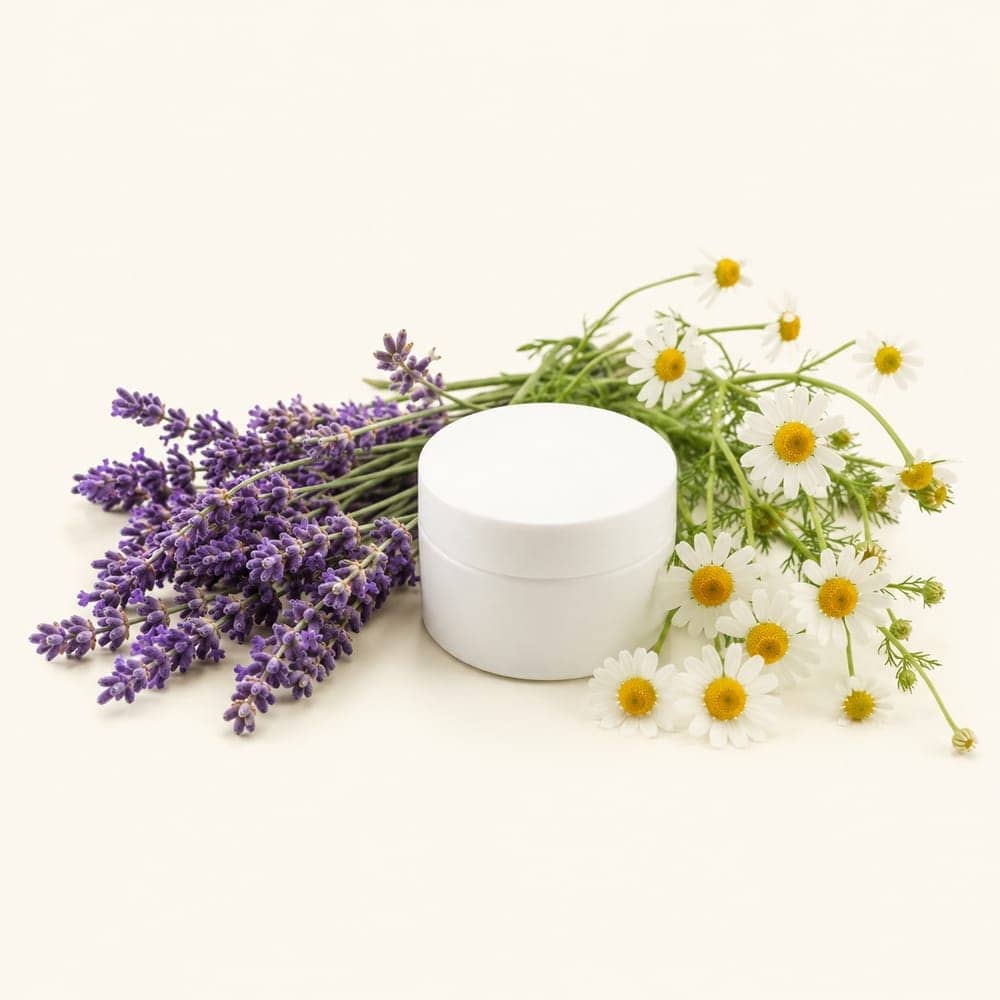 Sprigs of lavender and chamomile beside an unbranded cream jar on a seamless warm cream background.