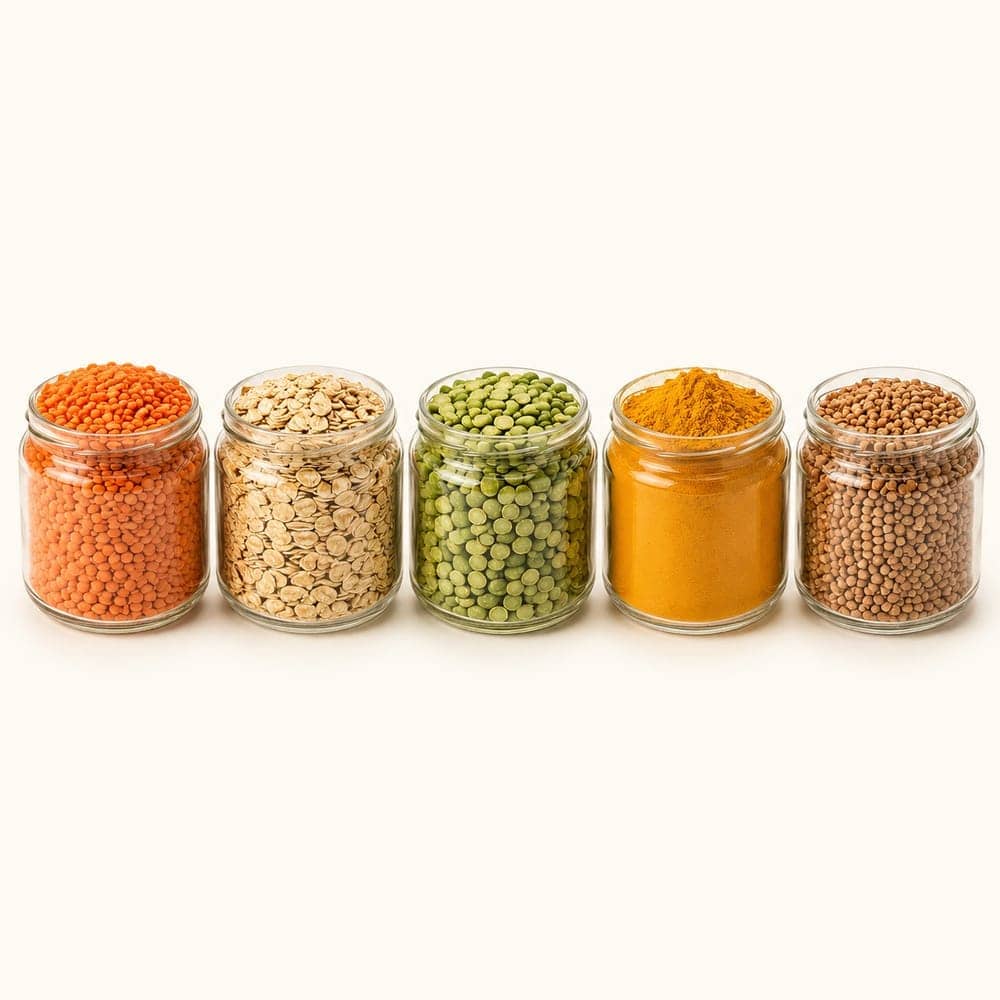 Five small glass jars of vivid raw ingredients — red lentils, golden oats, green peas, turmeric and buckwheat — on a seamless warm cream background.