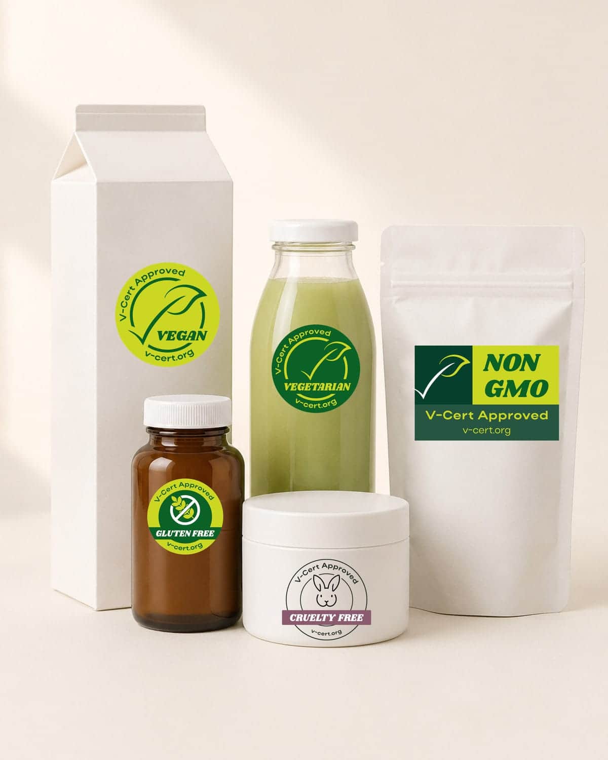 Five unbranded retail packages grouped on a warm cream background in bright daylight, each carrying one V-Cert mark: Vegan on a carton, Vegetarian on a bottle of green juice, Gluten-Free on an amber supplement bottle, Non-GMO on a stand-up pouch and Cruelty-Free on a cosmetic jar.